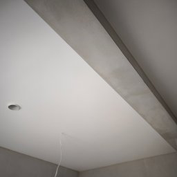 Buda-Mex Damian Bogucki - Gładki, biały sufit z zamontowanym punktowym oświetleniem i szarą belką konstrukcyjną. Widok z dołu, podkreślający precyzję wykończenia.