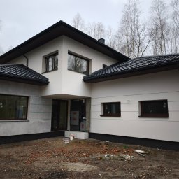 Buda-Mex Damian Bogucki - Nowoczesny dom jednorodzinny z białą elewacją i ciemną dachówką, częściowo wykończony, z widocznymi elementami przygotowania do dalszych prac.