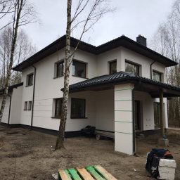 Buda-Mex Damian Bogucki - Dwukondygnacyjny dom z jasną elewacją i ciemnym dachem, otoczony drzewami. Widoczne detale wykończenia, takie jak rynny i okna. Ujęcie z poziomu gruntu.