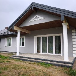 Buda-Mex Damian Bogucki - Szary dom jednorodzinny z białymi kolumnami i stolarką okienną, widok z zewnątrz na elewację i taras z kostki brukowej. Na dachu widoczne panele słoneczne.