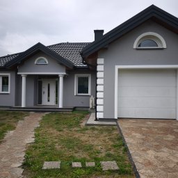 Buda-Mex Damian Bogucki - Szary dom jednorodzinny z białą stolarką i garażem, widok z ogrodu. Kamienna ścieżka prowadzi do wejścia. Dach pokryty szarą dachówką. Elewacja z dekoracyjnymi elementami.