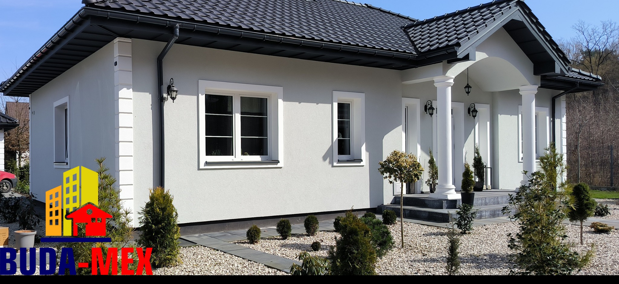 Nowoczesny dom jednorodzinny z jasną elewacją, ciemną dachówką i białymi kolumnami przy wejściu, otoczony ozdobnymi krzewami i kamieniami.