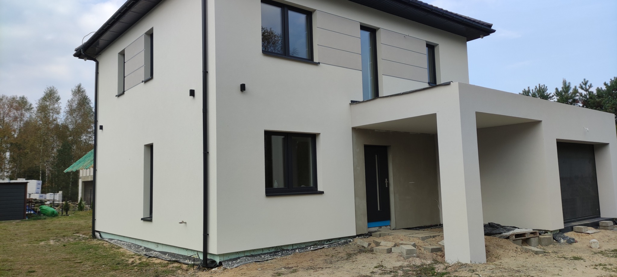 Nowoczesny dom z jasną elewacją, czarnymi oknami i elementami dekoracyjnymi, widok z zewnątrz w pochmurny dzień, na ziemi widoczne pozostałości po pracach budowlanych.