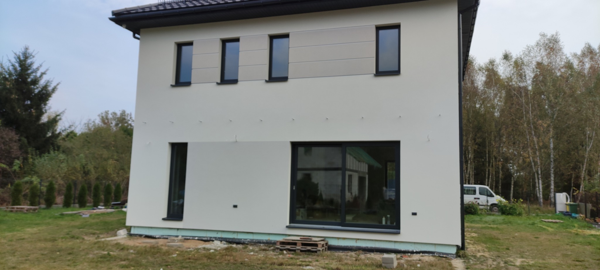 Nowoczesny dom z jasną elewacją, z fragmentem elewacji wykończonym panelami w odcieniach beżu, widoczne czarne ramy okienne i drzwi balkonowe, palety i styropian przy fundamencie.