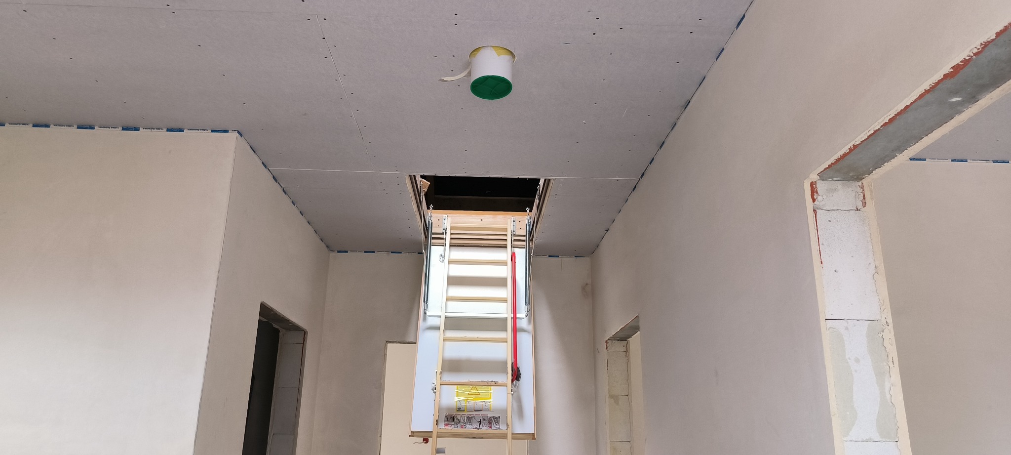 Wnętrze w trakcie remontu: ściany przygotowane do malowania, widoczne ślady szpachlowania, sufit z płyt gipsowo-kartonowych, rozkładane schody strychowe i otwór w suficie, taśmy malarskie...