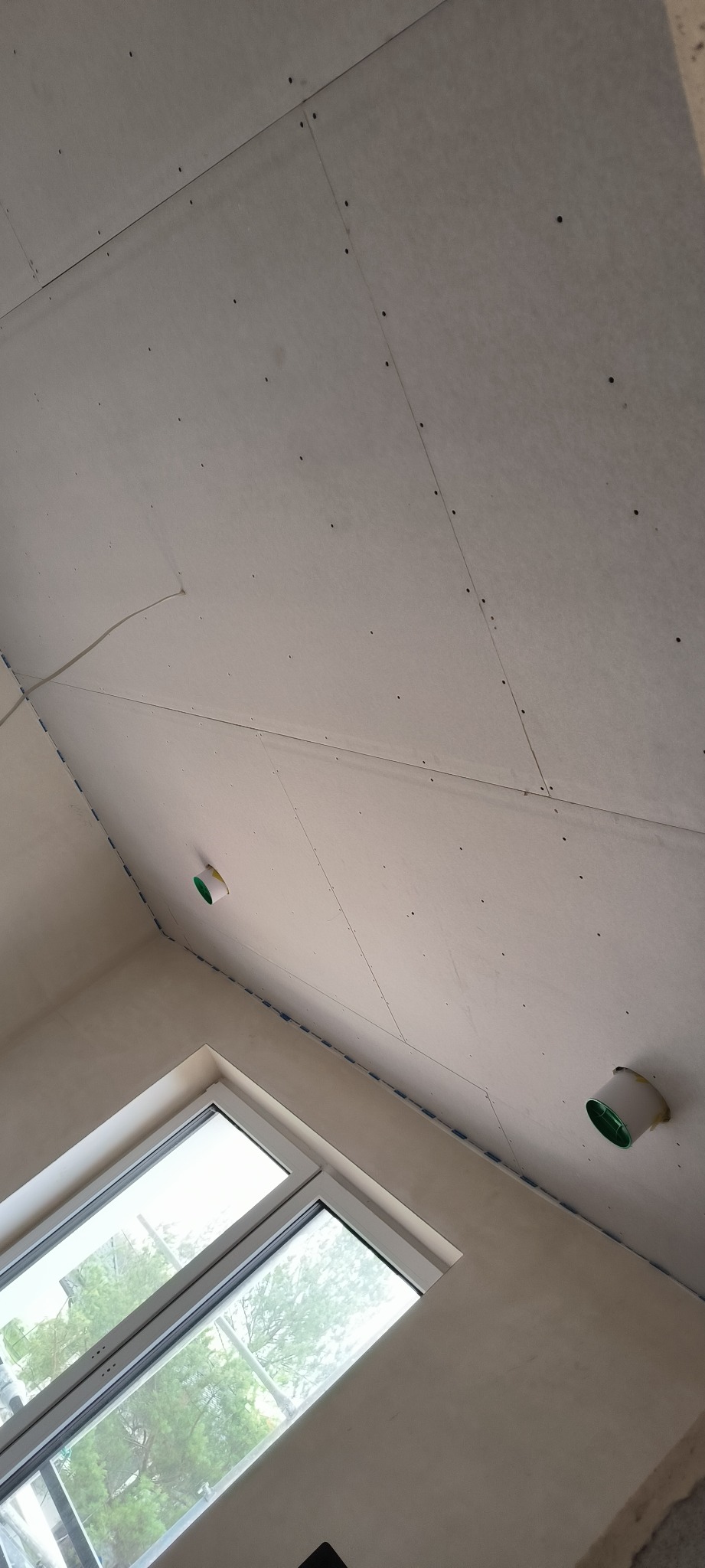 Poddasze w trakcie wykańczania płytami gipsowo-kartonowymi, widoczne wkręty, otwory wentylacyjne i okno dachowe z widokiem na drzewa.
