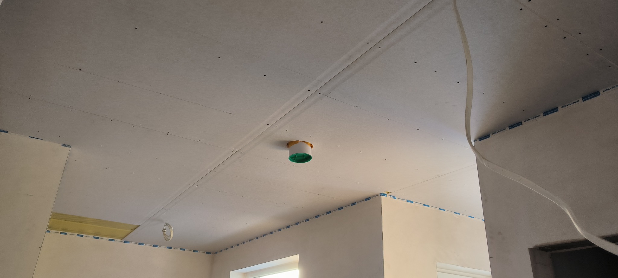 Wnętrze w trakcie montażu płyt gipsowo-kartonowych na suficie, widoczne łączenia płyt, taśma ślizgowa przy ścianach, otwór na oświetlenie oraz fragment ocieplenia wełną mineralną.