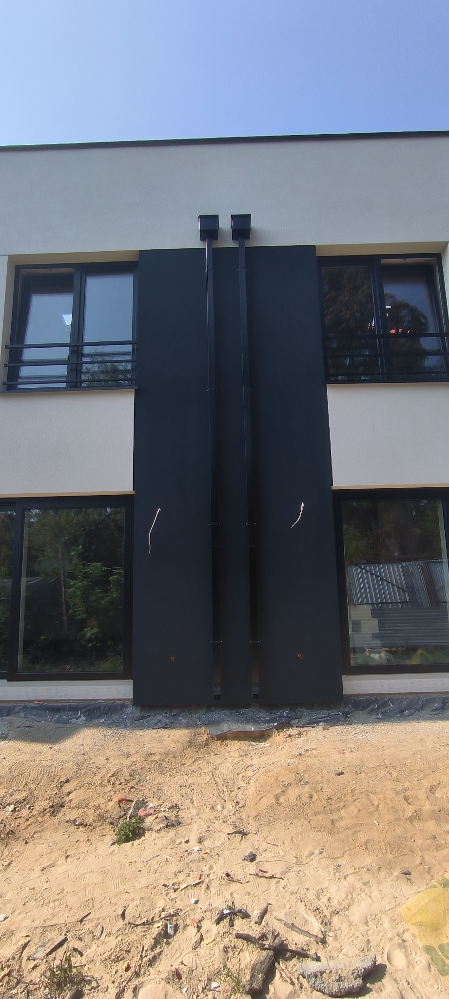 Nowoczesna elewacja budynku z ciemnymi panelami i jasnym tynkiem, widoczne okna z czarnymi ramami, teren wokół budynku w trakcie prac wykończeniowych.