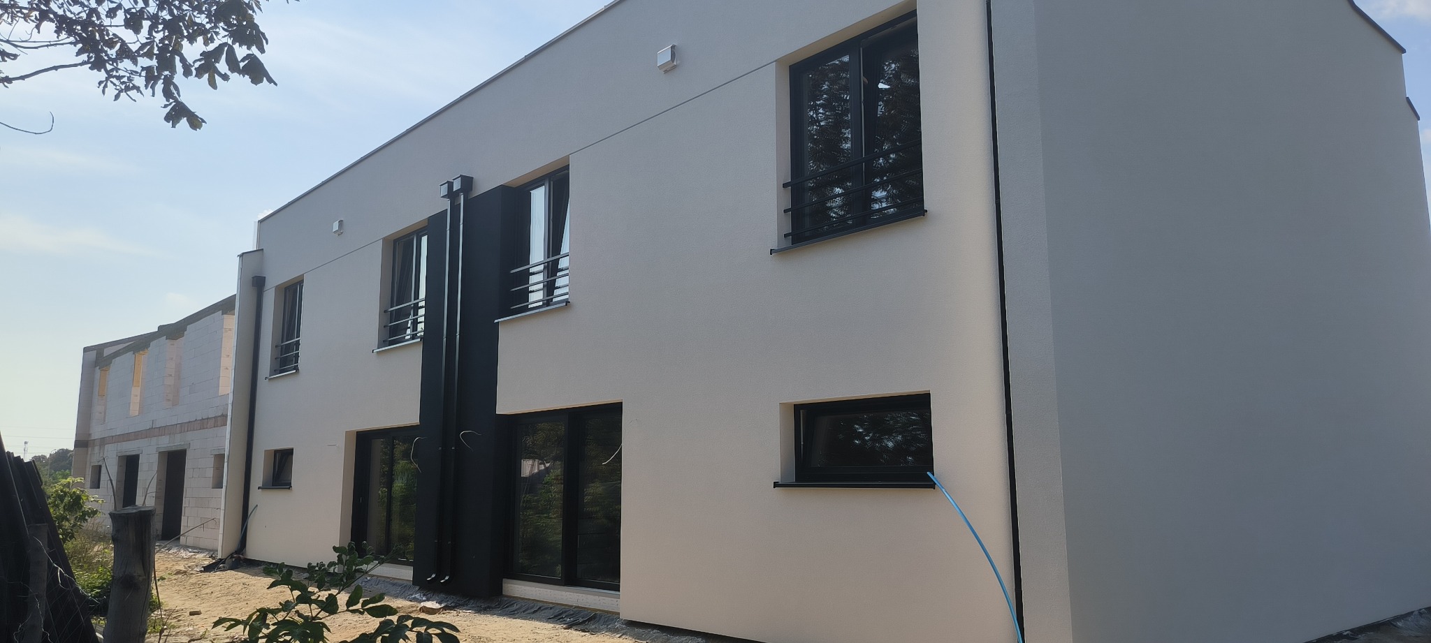 Nowoczesny dom z jasną elewacją i czarnymi oknami, widoczna instalacja odprowadzająca wodę deszczową, obok budynek w stanie surowym.