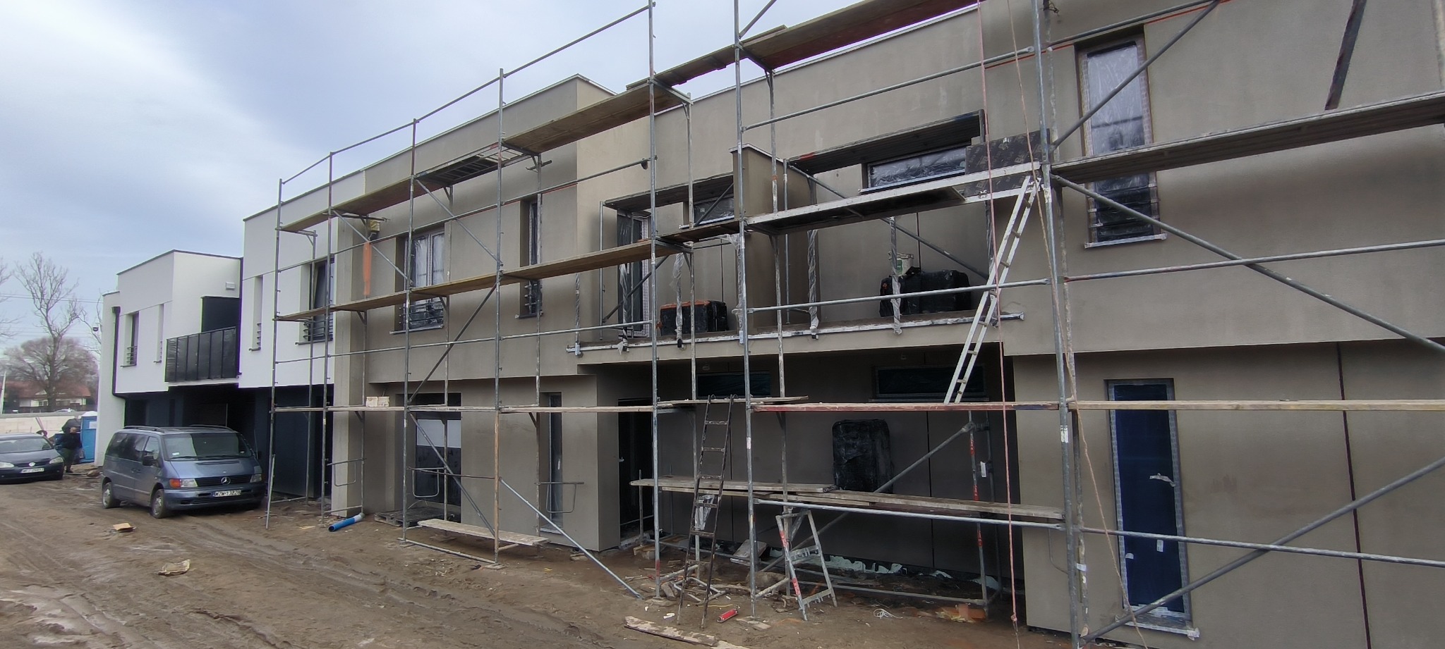 Szeregówka w trakcie wykonywania elewacji: budynek w szarym tynku, rusztowania, zabezpieczone okna folią, zaparkowany van.