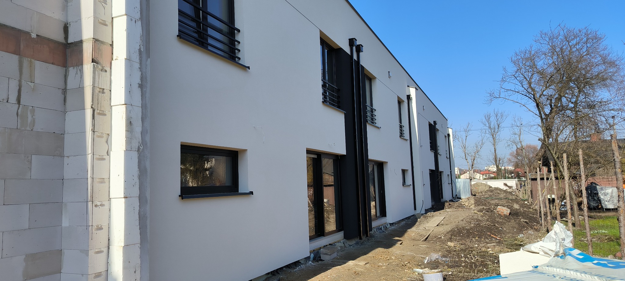 Nowoczesny budynek w trakcie wykańczania elewacji, widoczne fragmenty nieotynkowanych ścian z bloczków, biała, gładka elewacja, czarne obramowania okien i rur spustowych, teren budowy z resztkami...
