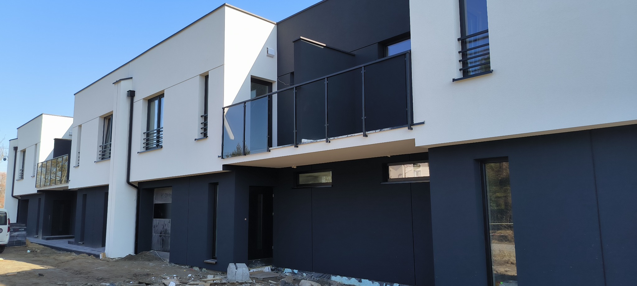 Nowoczesne szeregówki z elewacją w kolorze białym i grafitowym, balkony z czarnymi balustradami, teren wokół w trakcie prac wykończeniowych.