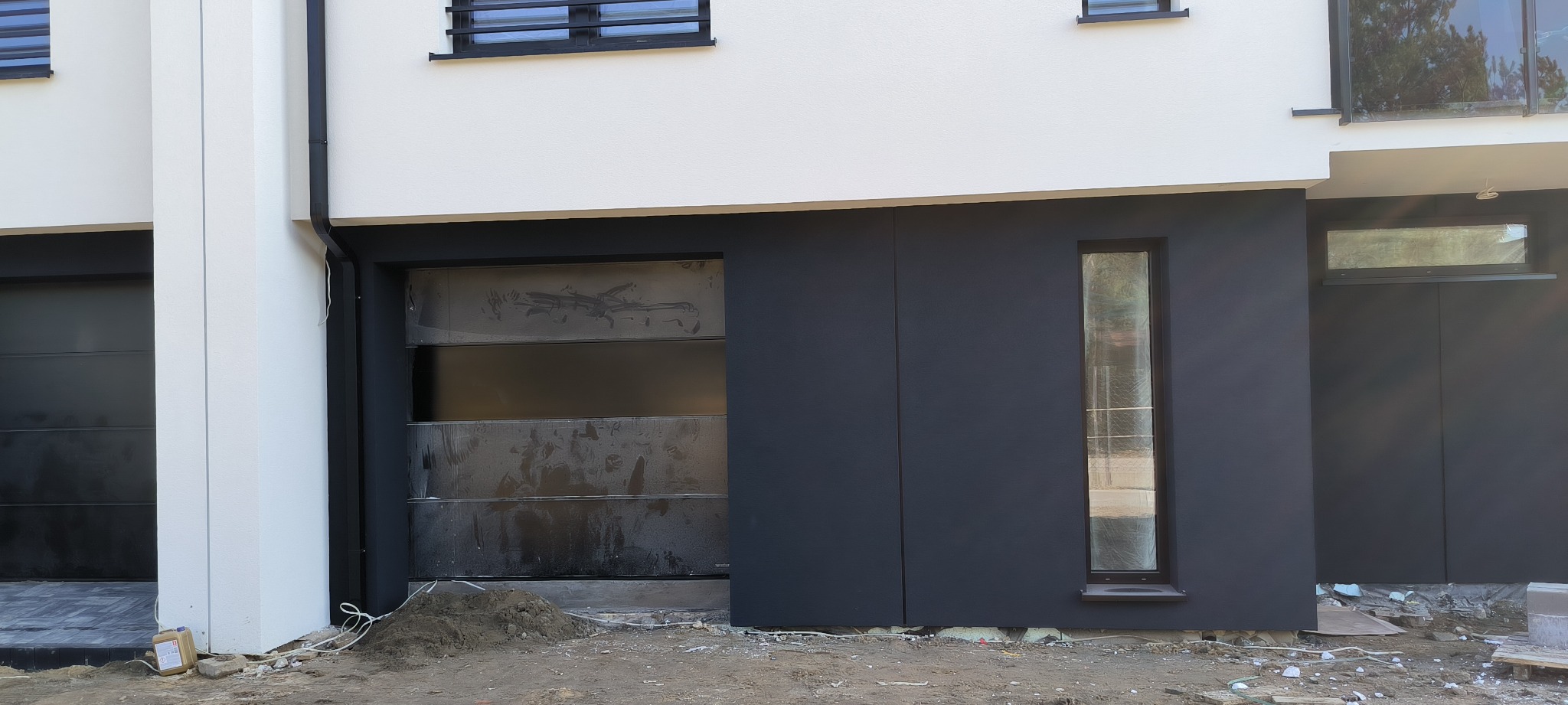 Nowoczesny dom w trakcie prac elewacyjnych: fragment fasady z szarą, gładką powierzchnią obok okna i garażu z folią ochronną, widoczne przygotowania do dalszych prac wykończeniowych.