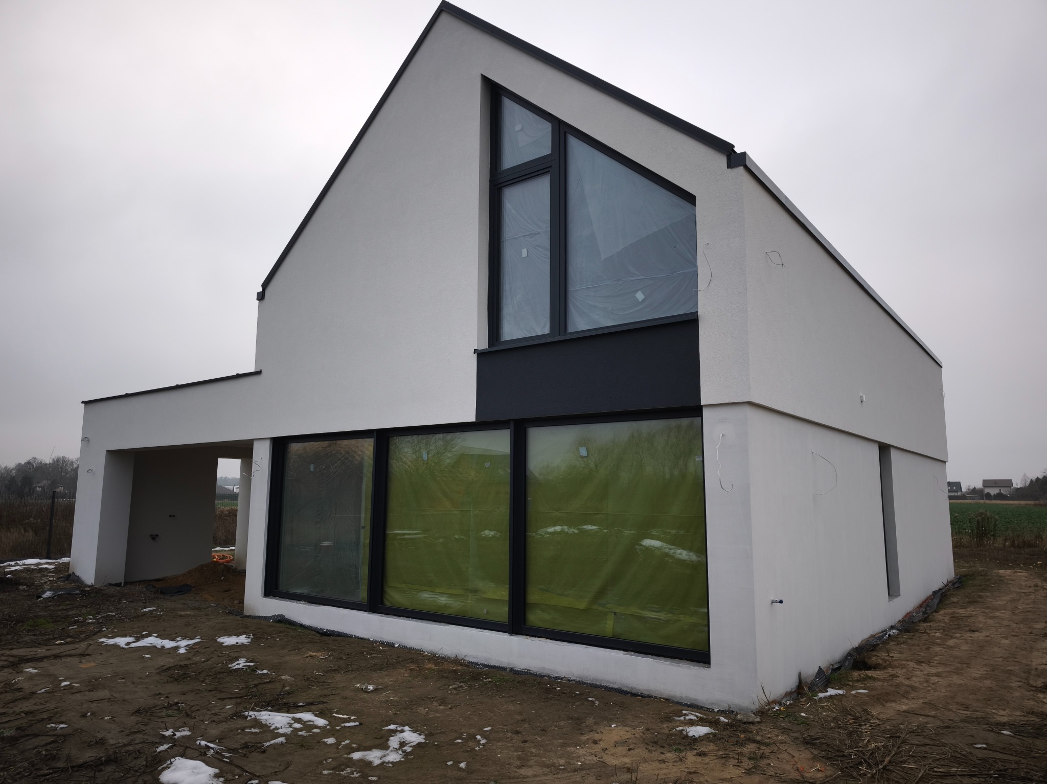 Nowoczesny dom z białą elewacją i dużymi oknami, częściowo zakrytymi folią ochronną. Minimalistyczny design, czarne ramy okienne, surowy krajobraz w tle.