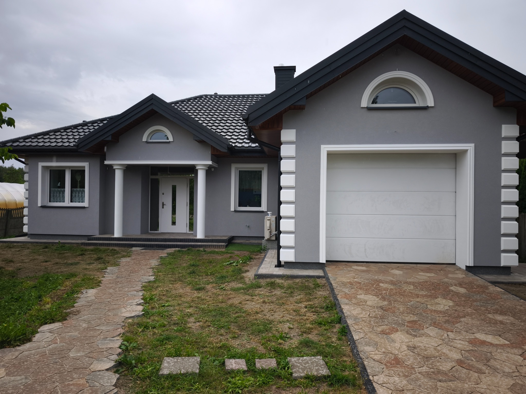 Szary dom jednorodzinny z białą stolarką okienną i garażem, widok z przodu. Kamienna ścieżka prowadzi do wejścia. Dach pokryty szarą dachówką.