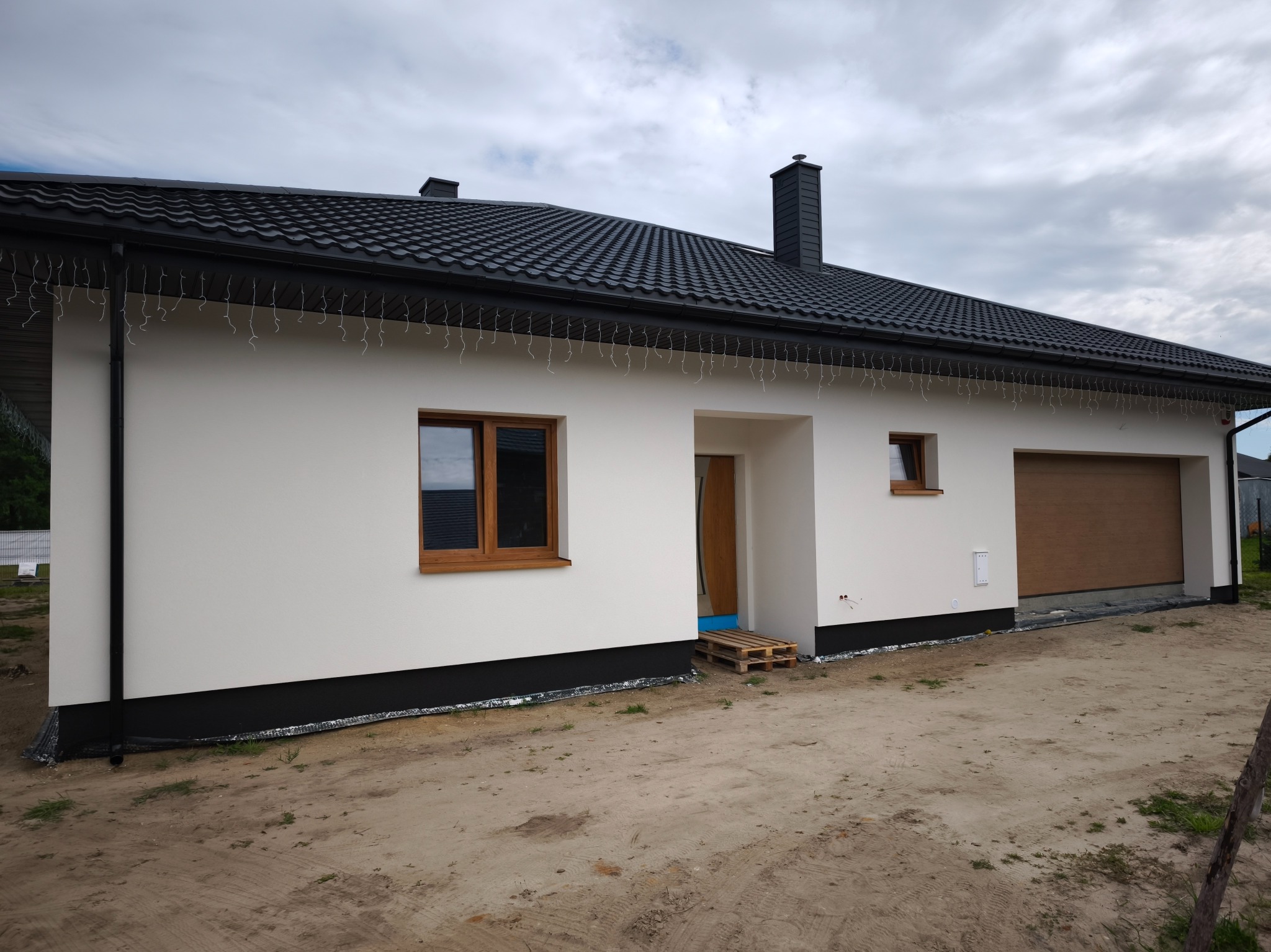 Biały dom parterowy z czarnym dachem i drewnianymi oknami. Widoczne detale wykończenia elewacji i otoczenie w trakcie prac ziemnych. Paleta przy wejściu.