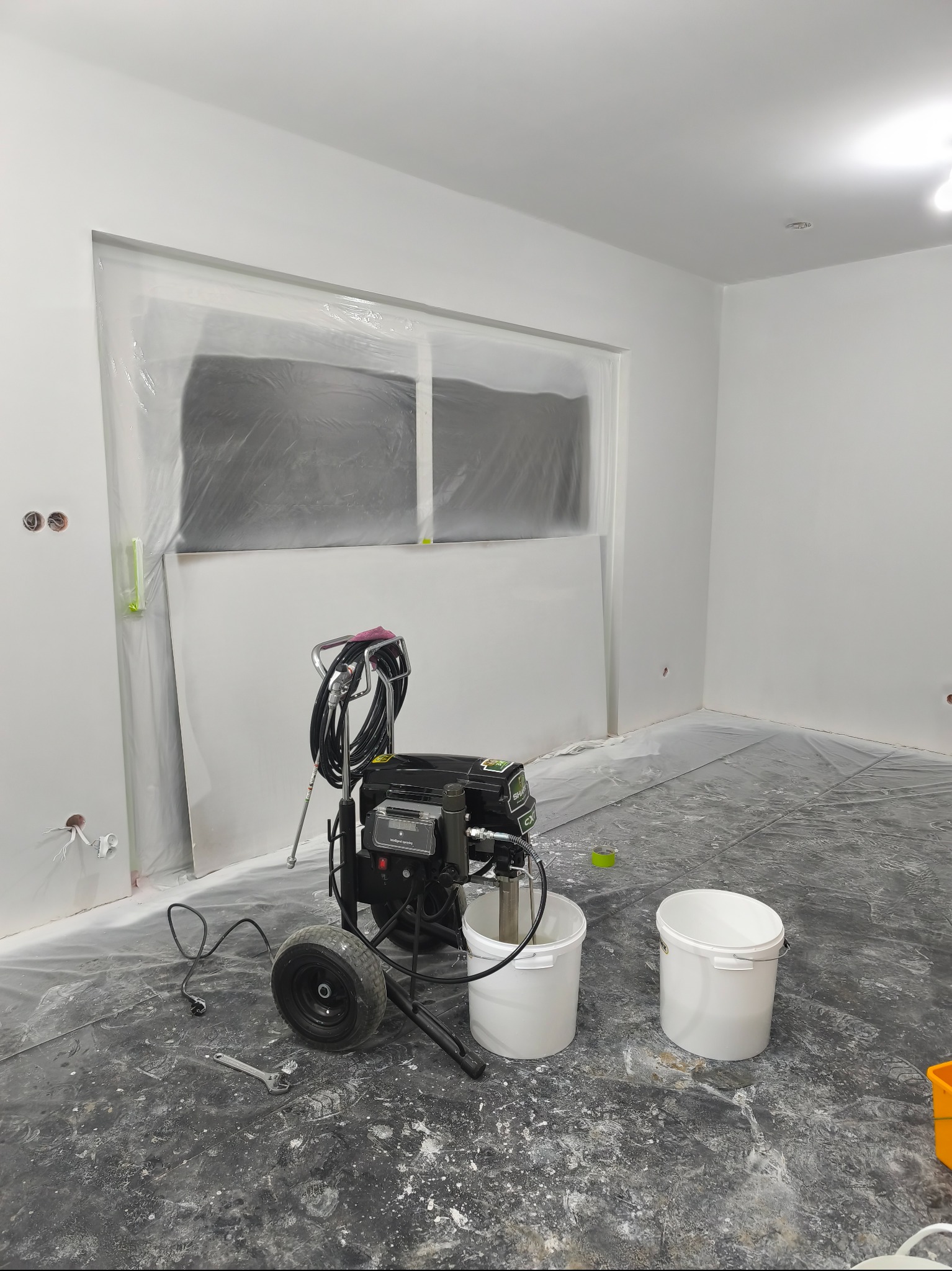 Wnętrze po malowaniu natryskowym: białe ściany, okno zabezpieczone folią, agregat malarski, puste wiadra i narzędzia na podłodze pokrytej folią ochronną.