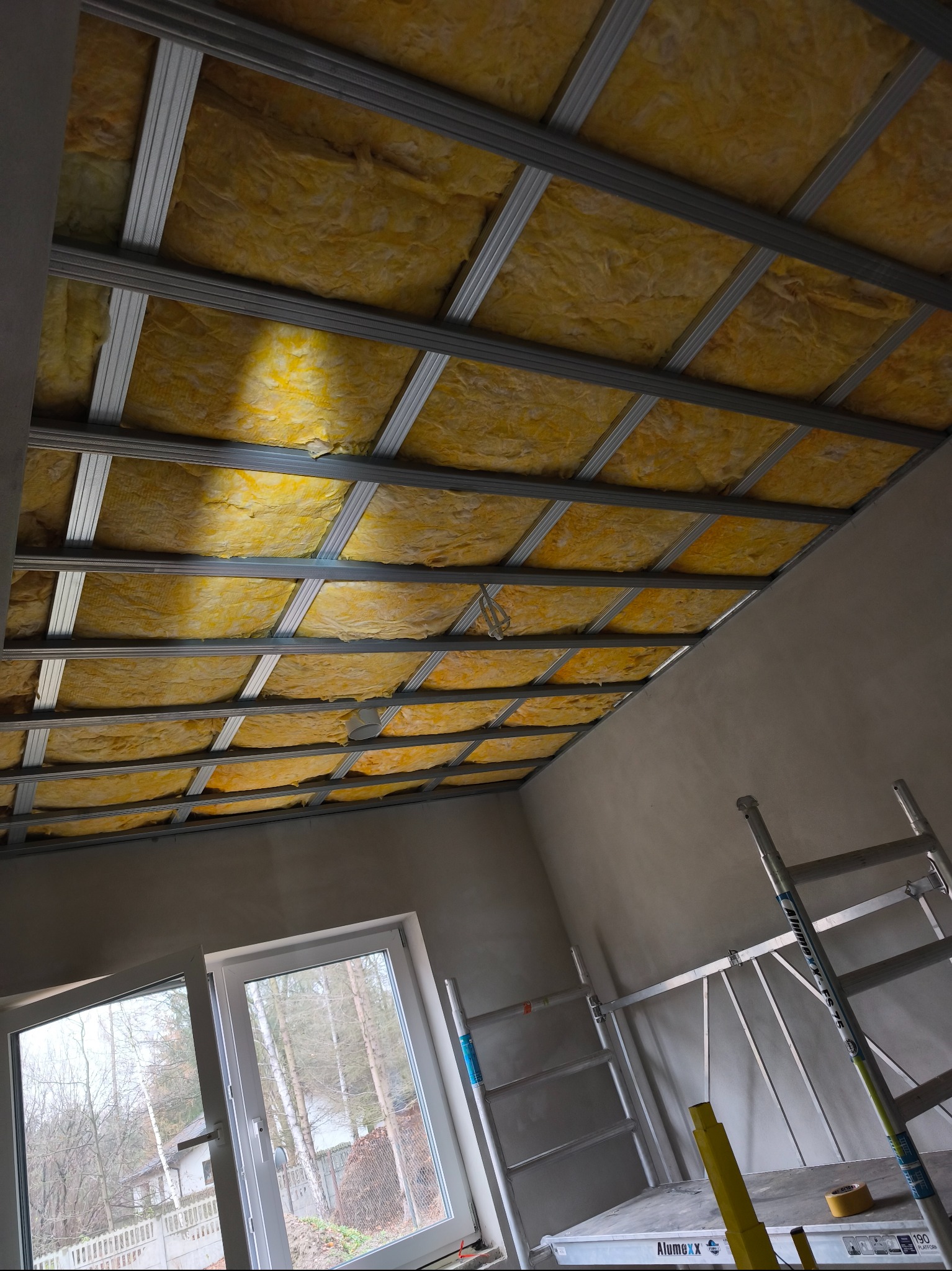 Montaż izolacji z wełny mineralnej w metalowym stelażu sufitu podwieszanego, widok z dołu, drabina w tle, okno z widokiem na drzewa.