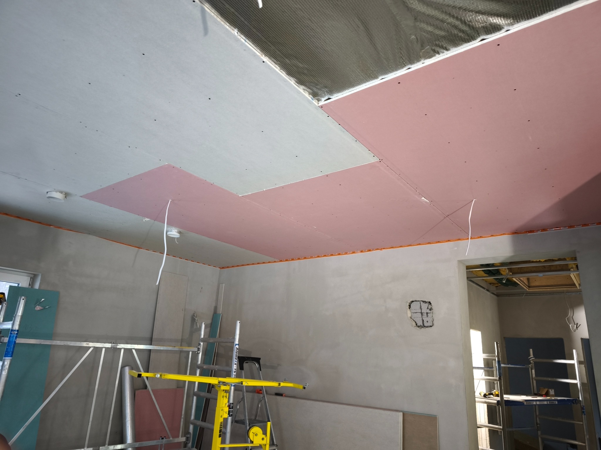 Montaż płyt karton-gips na suficie z widoczną izolacją. W tle ściany w stanie surowym i rusztowania. Widoczne kable i otwór w ścianie.