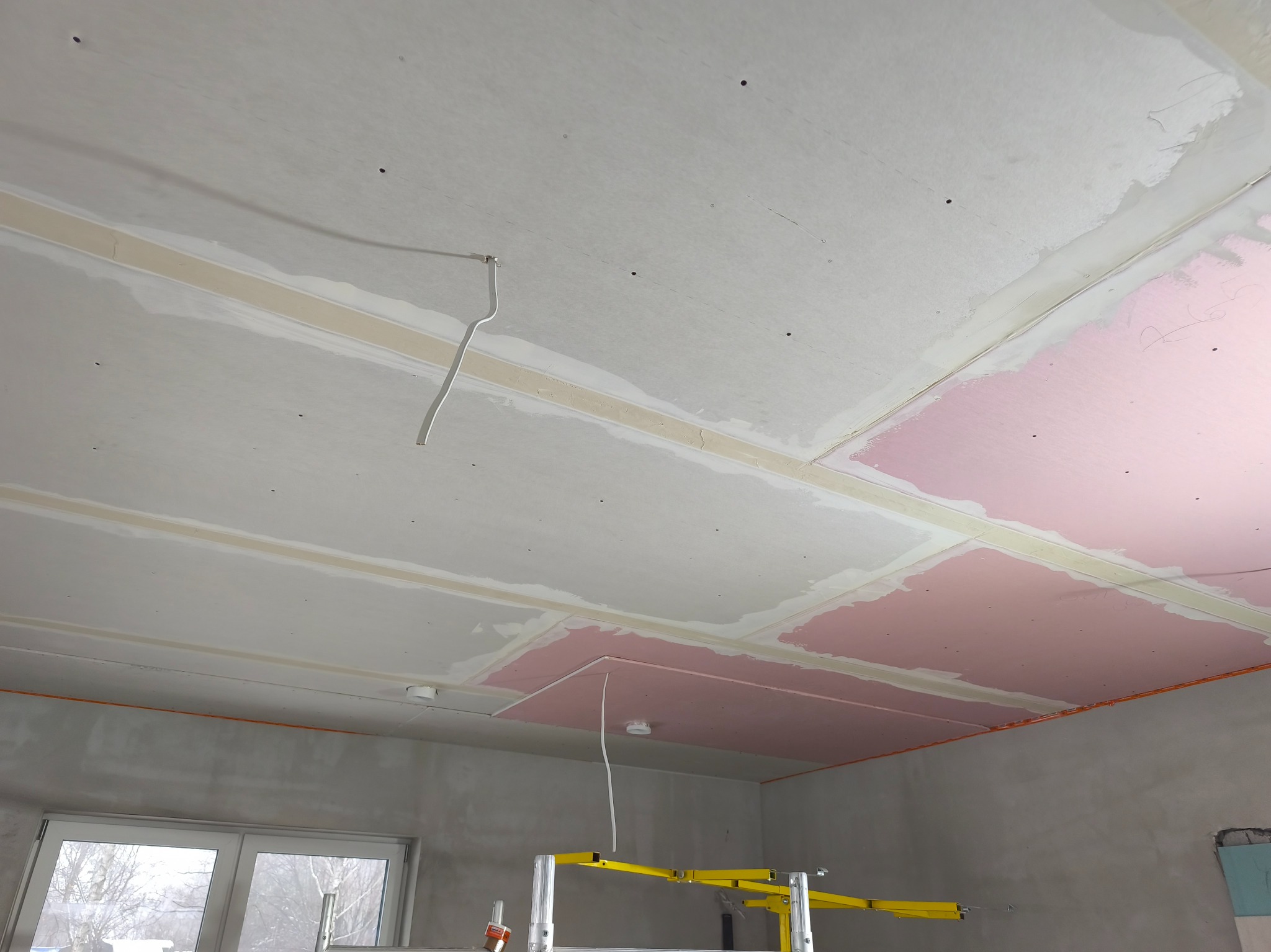 Montaż płyt gipsowo-kartonowych na suficie, widoczne łączenia i fragmenty różowej płyty. W tle ściana i rusztowanie malarskie.