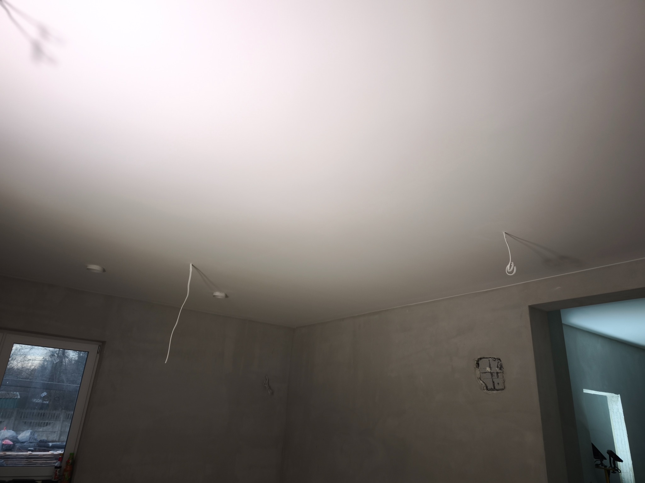 Biały sufit napinany z wystającymi kablami oświetleniowymi w szpachlowanym pomieszczeniu. Widoczne okno i fragment przejścia do innego pokoju.