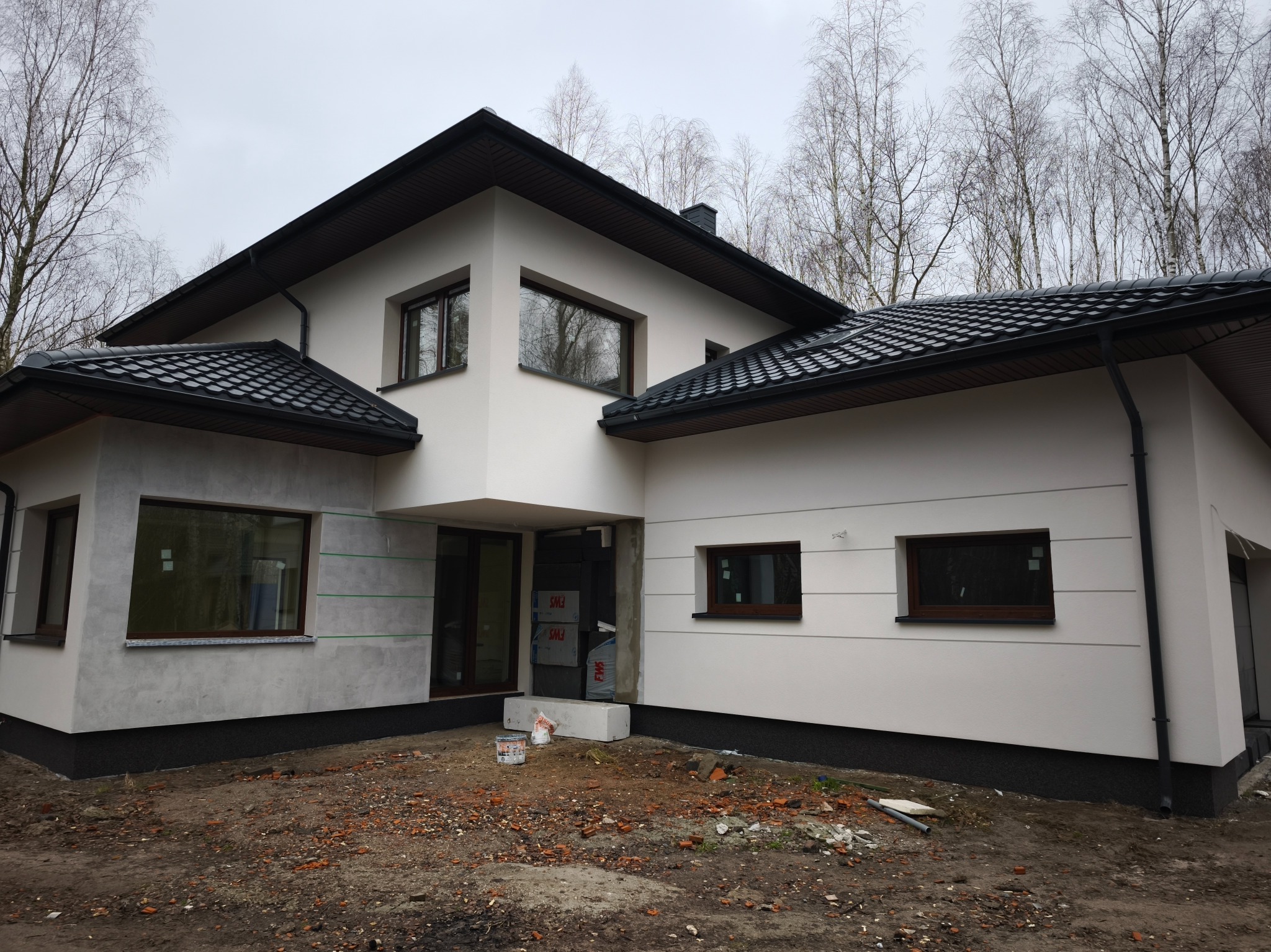 Nowoczesny dom jednorodzinny z białą elewacją i ciemną dachówką, częściowo wykończony, z widocznymi elementami przygotowania do dalszych prac.