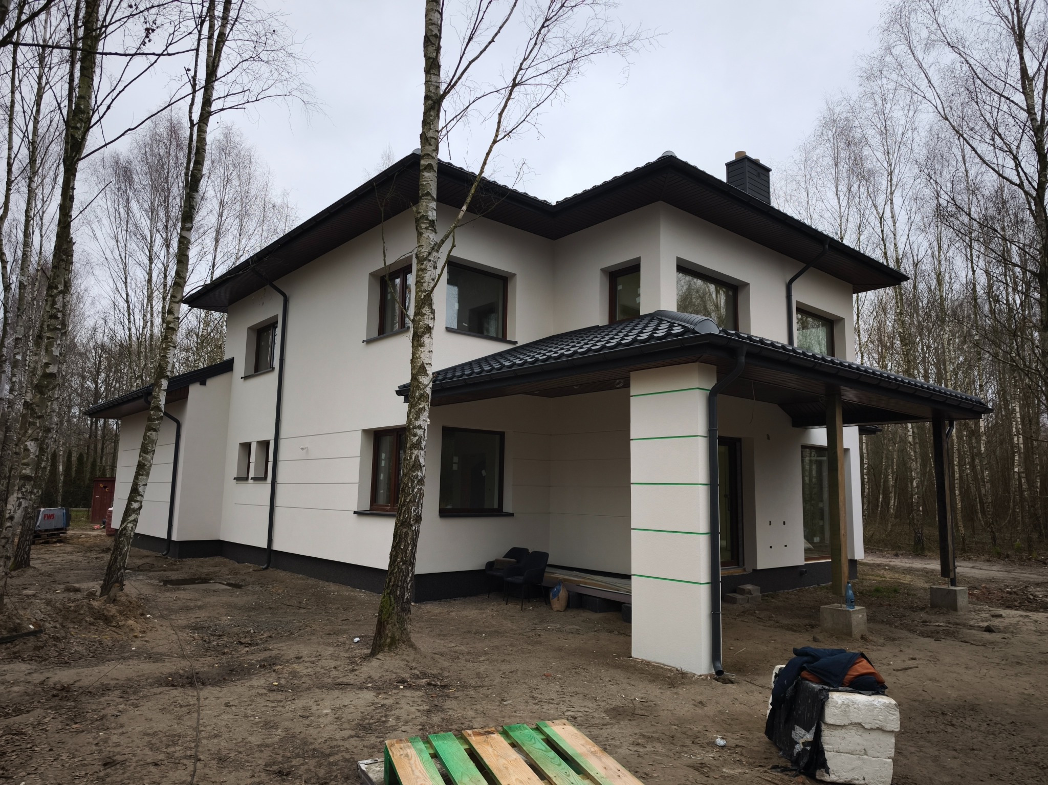 Dwukondygnacyjny dom z jasną elewacją i ciemnym dachem, otoczony drzewami. Widoczne detale wykończenia, takie jak rynny i okna. Ujęcie z poziomu gruntu.