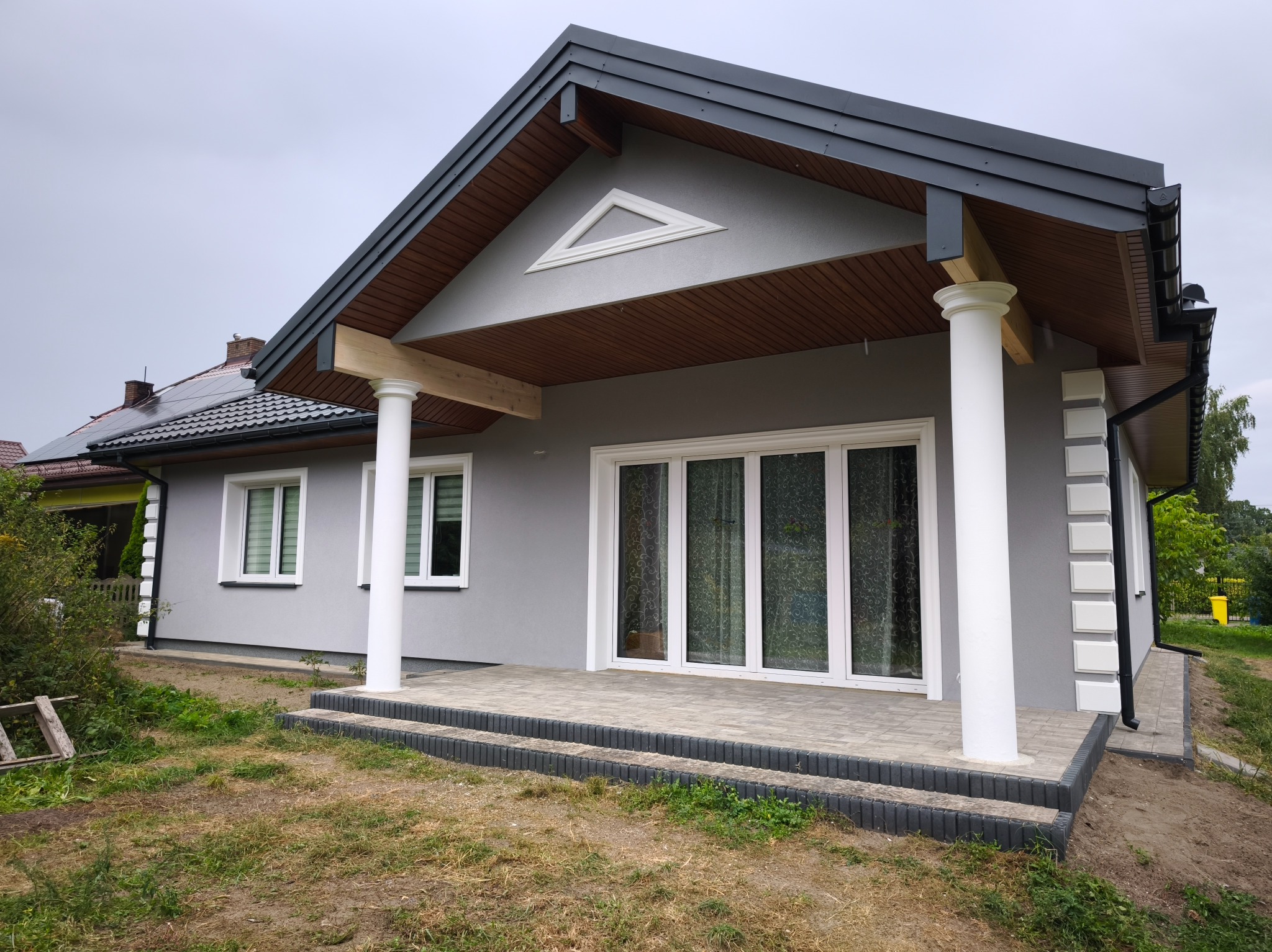 Szary dom jednorodzinny z białymi kolumnami i stolarką okienną, widok z zewnątrz na elewację i taras z kostki brukowej. Na dachu widoczne panele słoneczne.