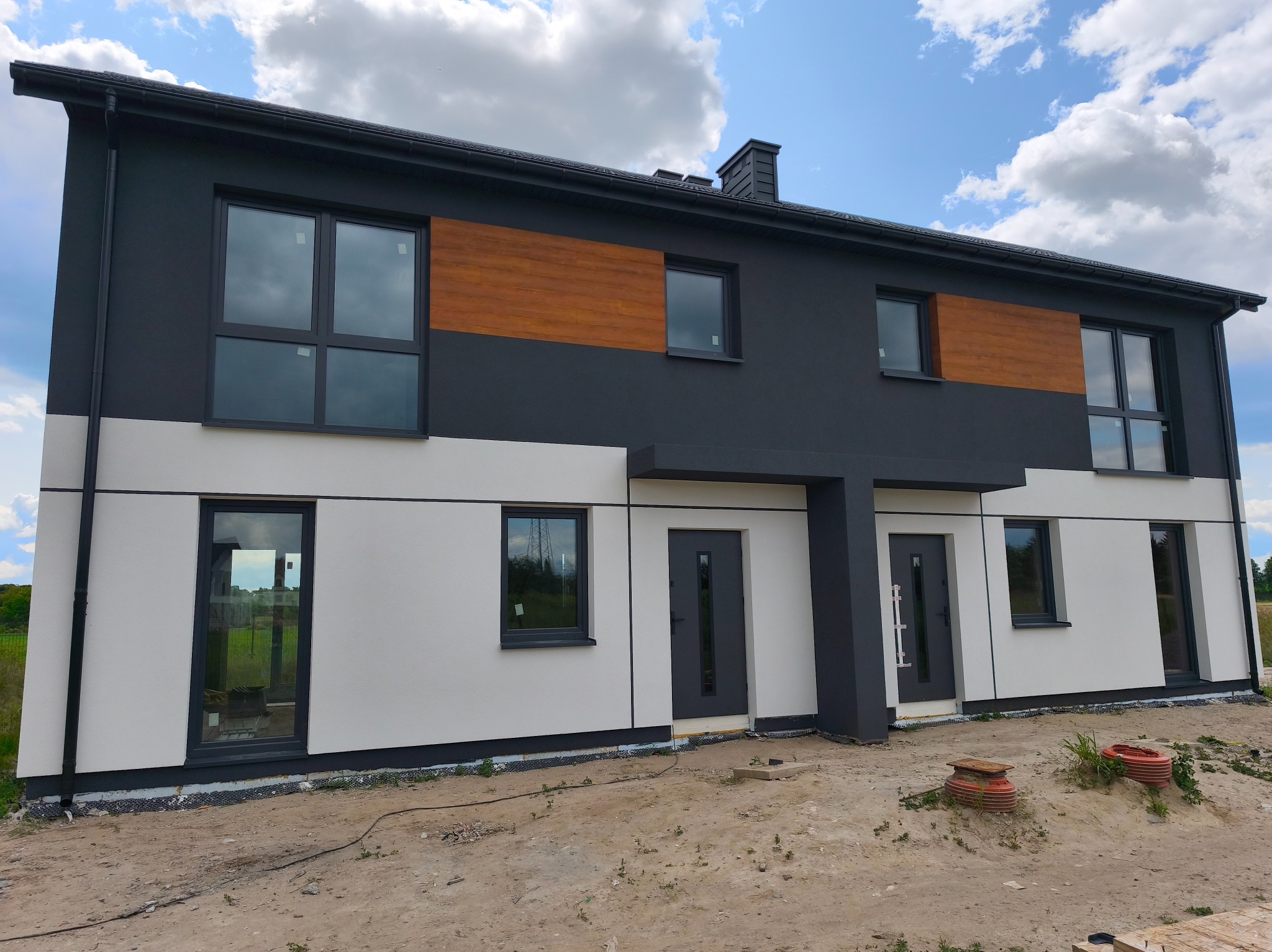 Dwukondygnacyjny dom bliźniak z elewacją w odcieniach szarości i bieli, z drewnianym akcentem. Widoczne okna z ciemnymi ramami i dwie pary drzwi wejściowych.