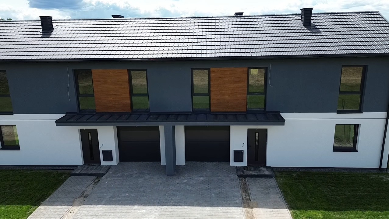 Dwukondygnacyjny dom bliźniak z szarą elewacją, drewnianymi akcentami i czarną dachówką. Widoczne dwa garaże z bramami segmentowymi i chodnik z kostki.