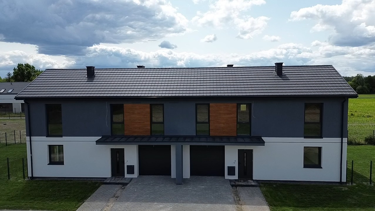 Dwukondygnacyjny bliźniak z szarą elewacją i drewnianymi panelami, widok z góry na tle pochmurnego nieba i zielonego pola. Nowoczesny design.