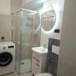 Nowoczesna łazienka z kabiną prysznicową, pralką, umywalką z szafką i okrągłym lustrem z podświetleniem LED, wykończona w szarościach.