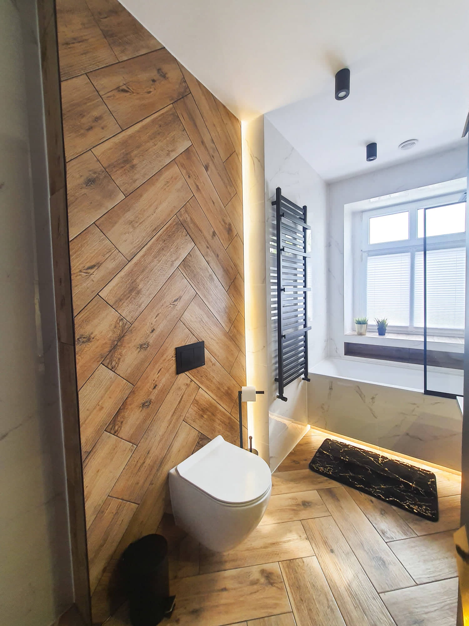 Nowoczesna łazienka z drewnopodobnymi płytkami ułożonymi w jodełkę na ścianie i podłodze, podświetleniem LED, czarnym grzejnikiem drabinkowym i wiszącą toaletą.