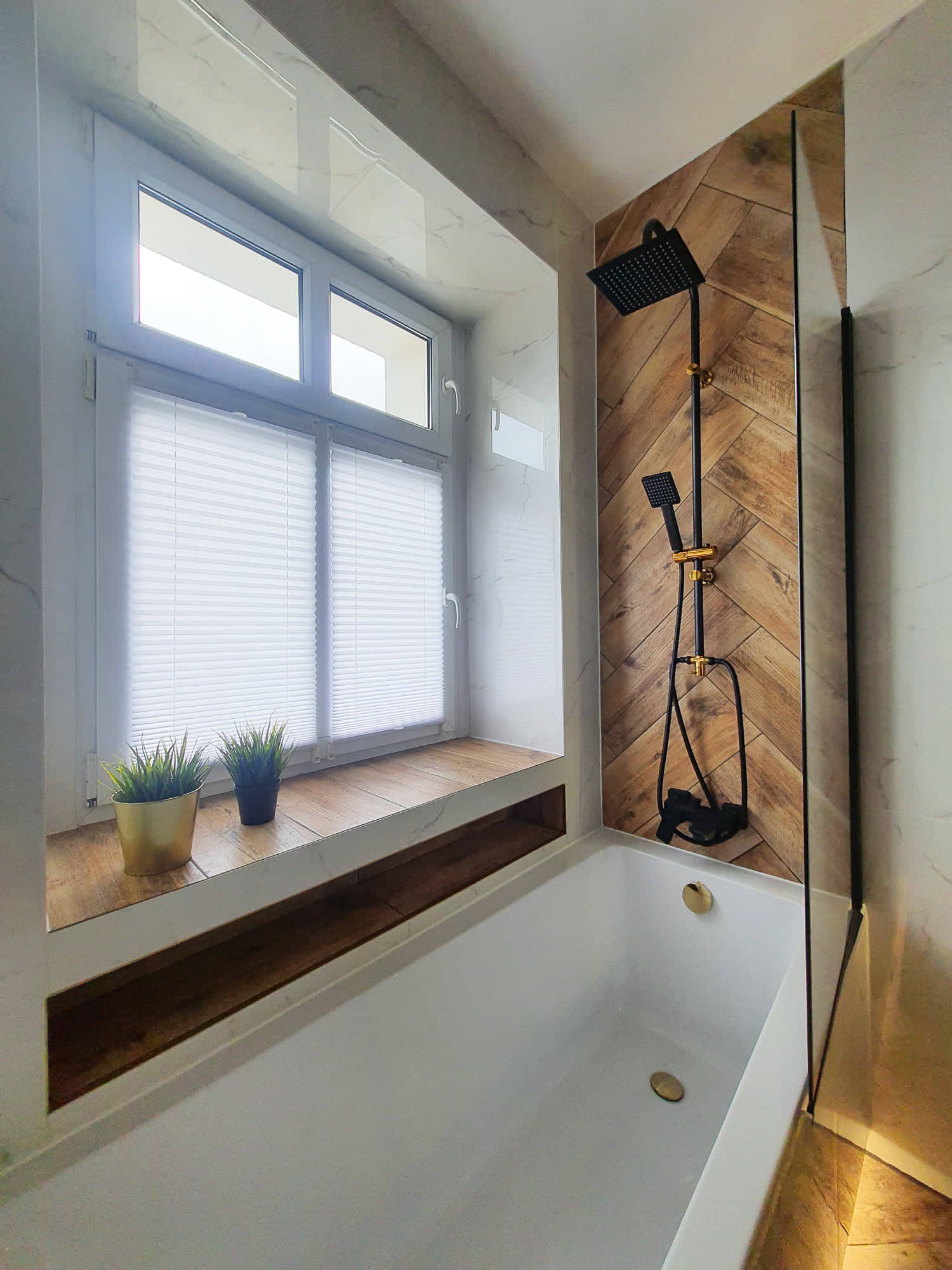 Nowoczesna łazienka z białą wanną, czarną armaturą prysznicową na tle ściany wyłożonej drewnopodobnymi płytkami ułożonymi w jodełkę, okno z żaluzjami plisowanymi i drewnianym parapetem z doniczkami.