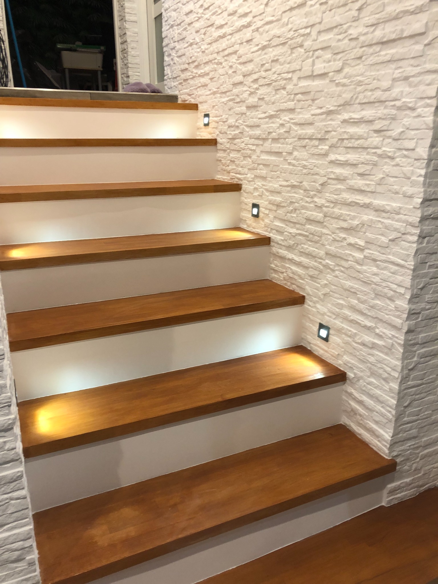 Nowoczesne schody z drewnianymi stopniami i białymi podstopnicami, oświetlone punktowymi lampkami w ścianie z białej cegły dekoracyjnej.