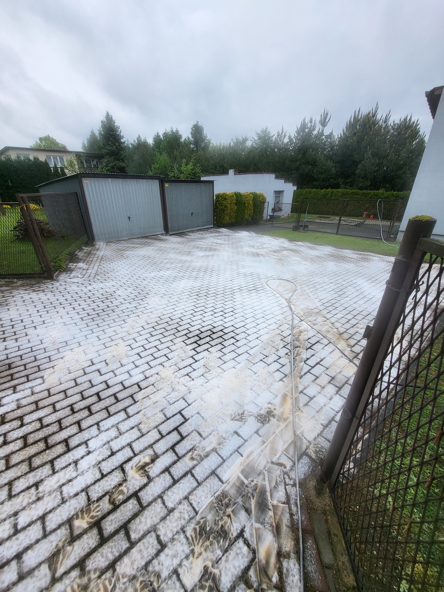 Częściowo umyta kostka brukowa na podjeździe, widoczne ślady po myciu ciśnieniowym, w tle garaże i zieleń.