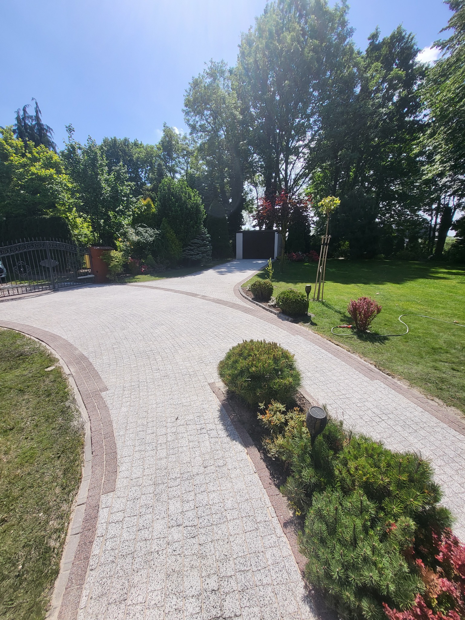 Szeroki, wybrukowany podjazd z jasnej kostki z ciemniejszym obramowaniem, prowadzący do posesji z bramą i zadbanym trawnikiem z krzewami.