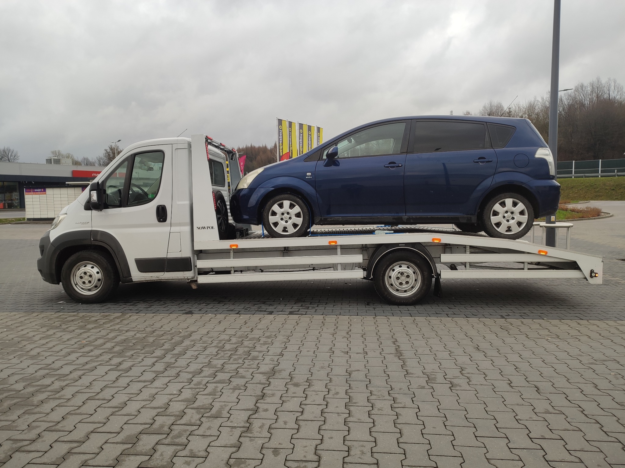 Biała laweta z niebieskim samochodem osobowym marki Toyota na platformie, zaparkowana na szarym brukowanym placu w pochmurny dzień.