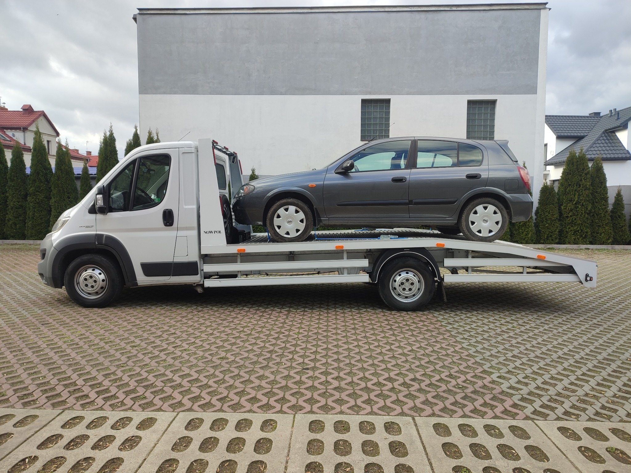 Szary samochód osobowy Nissan Almera na białej lawecie w trakcie transportu, na tle budynku i kostki brukowej.