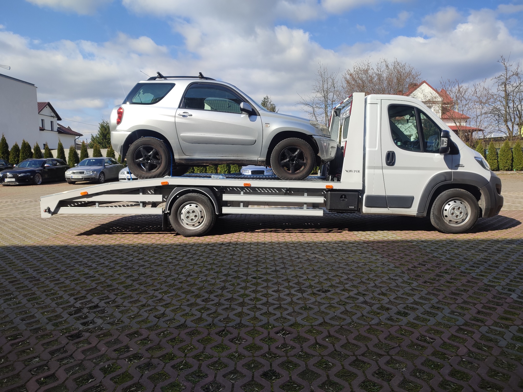 Srebrny samochód osobowy typu SUV przewożony na lawecie na tle budynków w Staszowie, widok z boku, w słoneczny dzień.