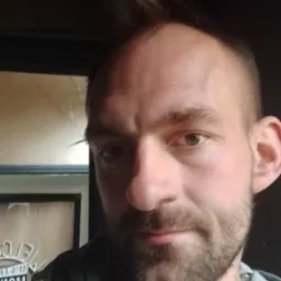 Portret mężczyzny z krótką brodą i pojedynczym kosmykiem włosów sterczącym do góry, ubrany w bluzę z kapturem na tle ciemnego pomieszczenia.