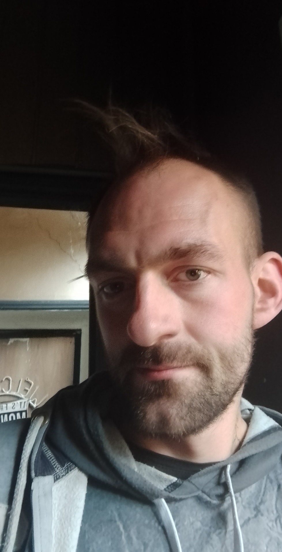 Portret mężczyzny z krótką brodą i pojedynczym kosmykiem włosów sterczącym do góry, ubrany w bluzę z kapturem na tle ciemnego pomieszczenia.