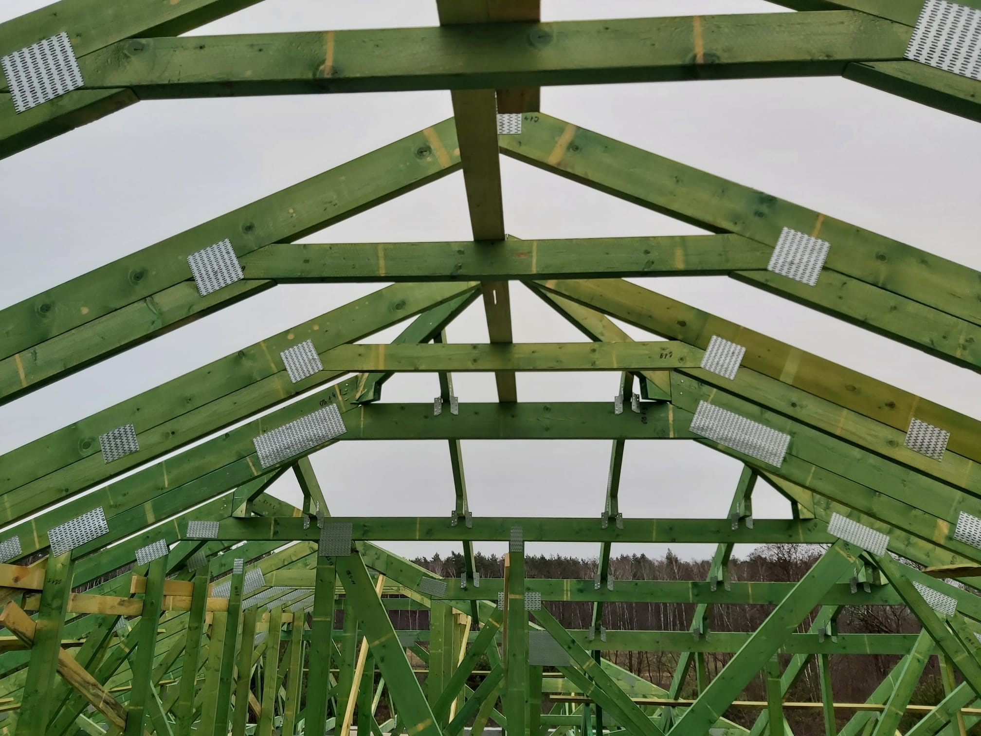 Wnętrze konstrukcji dachu z impregnowanego drewna, połączonego metalowymi płytkami perforowanymi, widok od dołu na tle szarego nieba i zalesionego horyzontu.