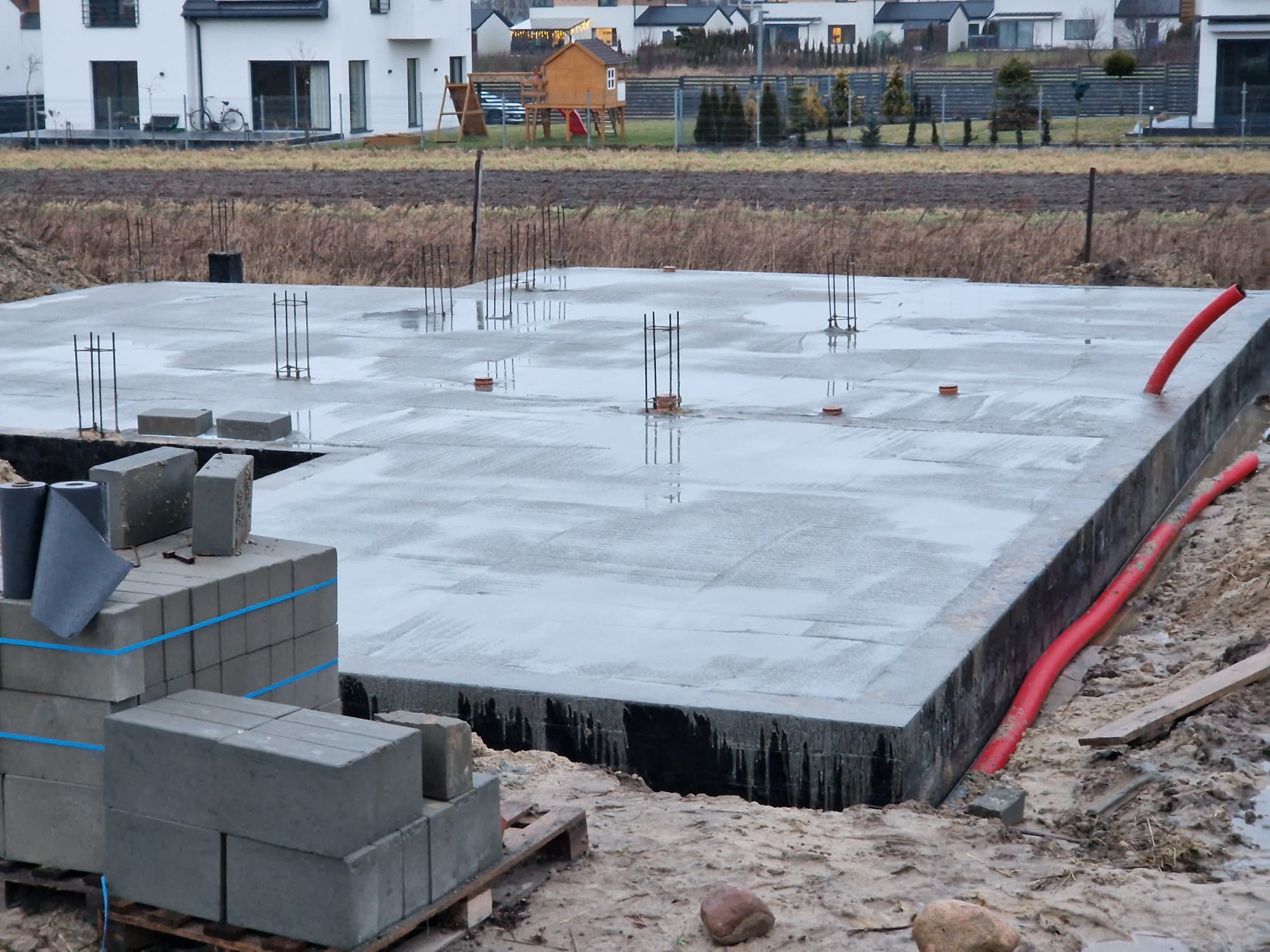 Wylana, mokra płyta fundamentowa z wystającymi prętami zbrojeniowymi, obok paleta z bloczkami betonowymi, w tle domy jednorodzinne w zabudowie szeregowej.