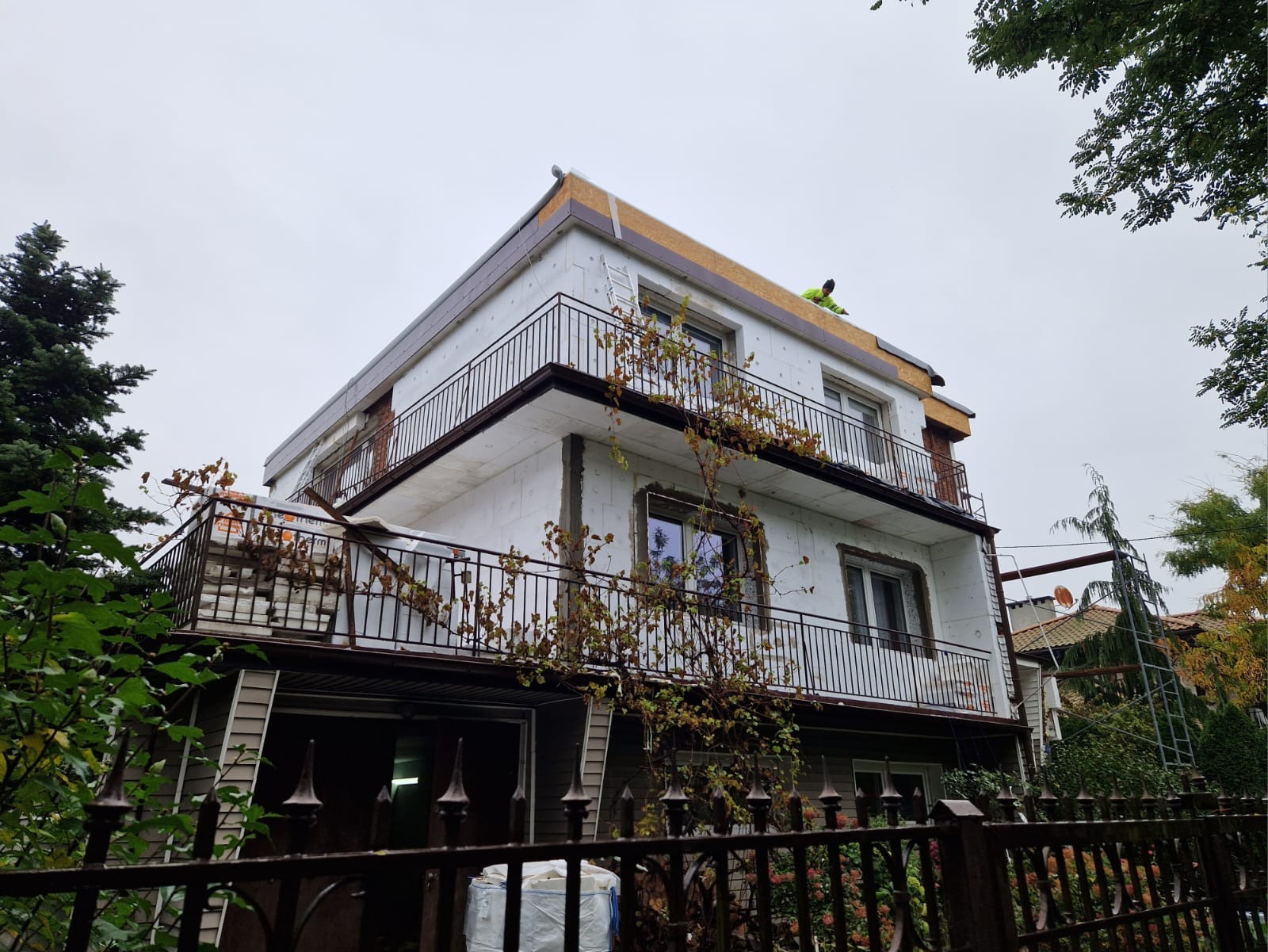 Dwupiętrowy dom w trakcie ocieplania elewacji styropianem, widoczny pracownik na dachu, zabezpieczone okna, balkon z metalową balustradą, widok zza ozdobnego ogrodzenia.
