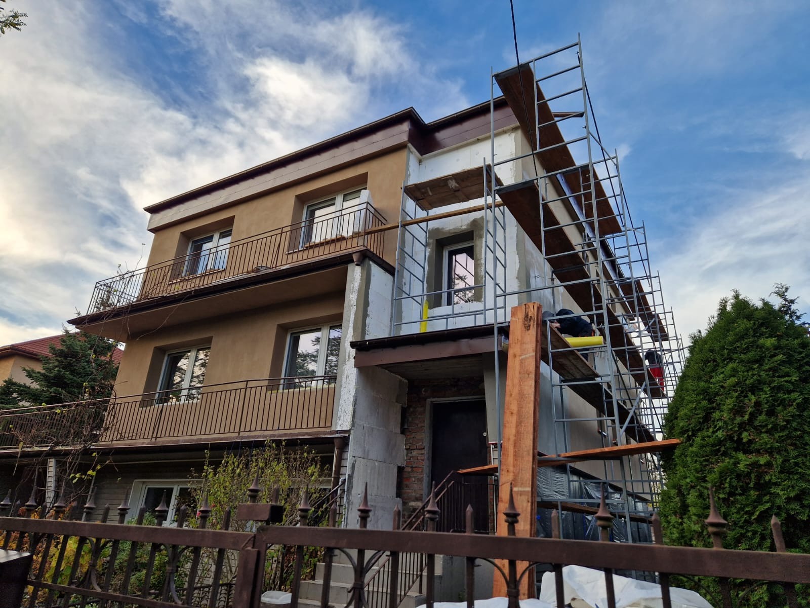 Dwupiętrowy dom w trakcie prac elewacyjnych, widoczne rusztowania i częściowo ocieplona ściana z białym styropianem, na tle błękitnego nieba z chmurami.