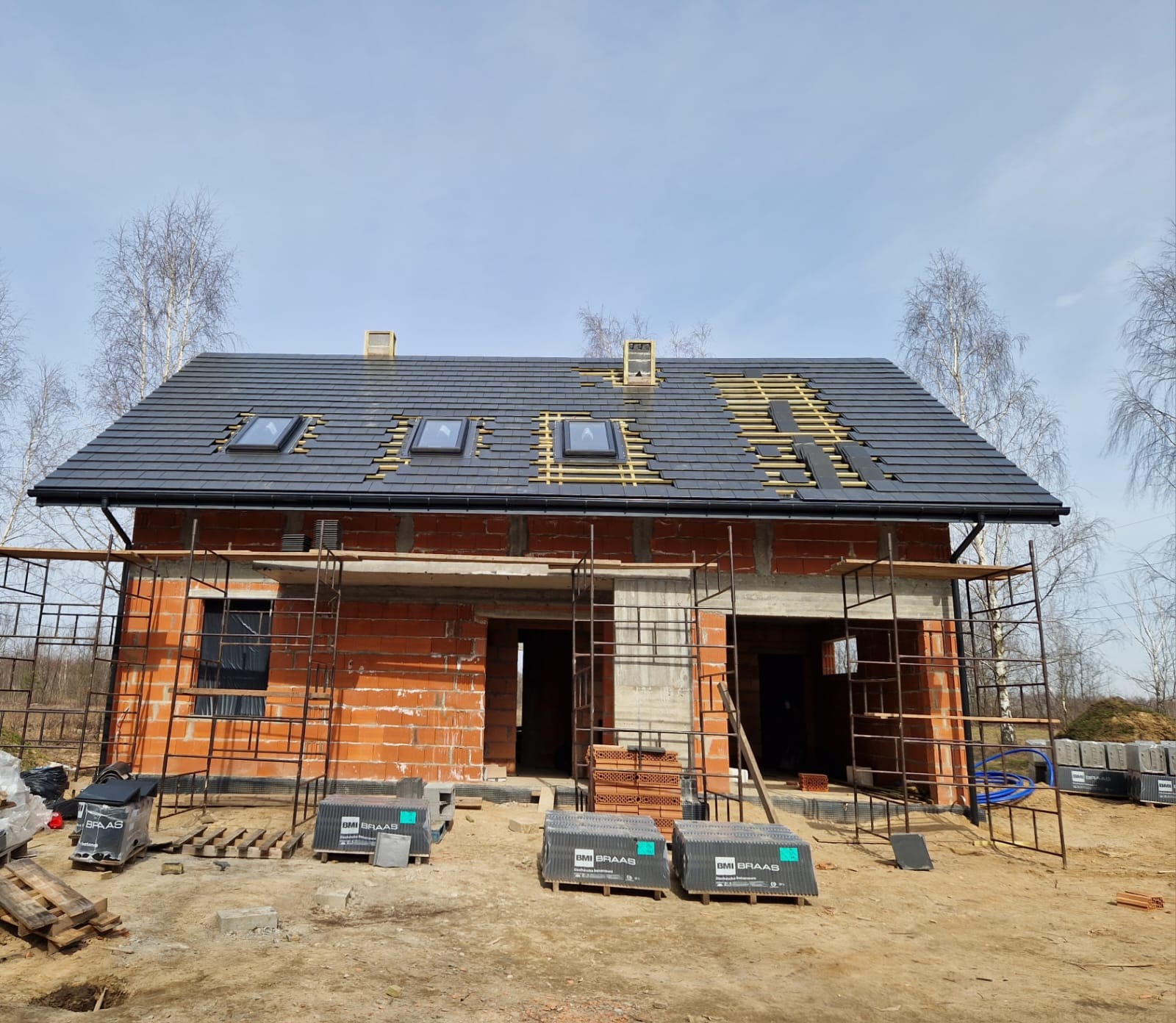 Wznoszony dom jednorodzinny z częściowo ułożonym, ciemnoszarym dachem z dachówką, widoczne okna dachowe i fragmenty konstrukcji drewnianej, na pierwszym planie palety z dachówką BMI Braas...
