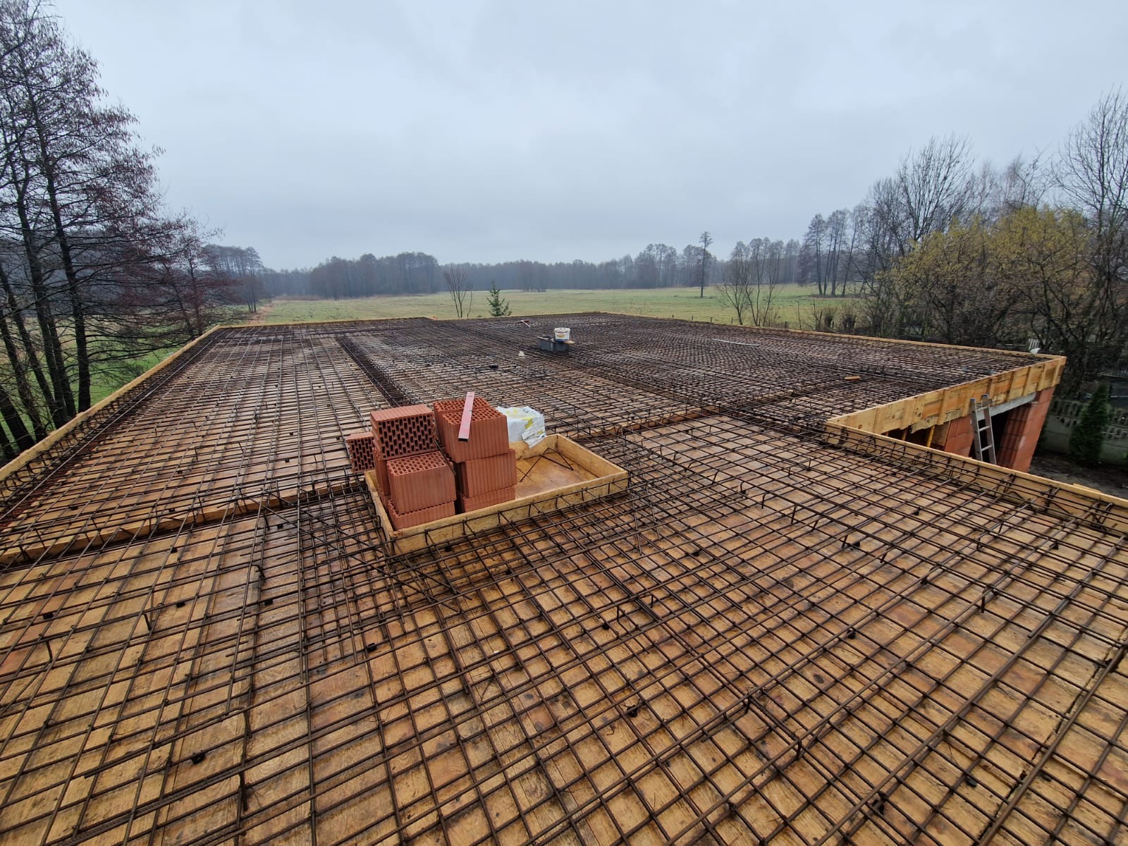 Zbrojenie płyty fundamentowej na deskowaniu, z widocznymi cegłami i krajobrazem w tle, w pochmurny dzień.