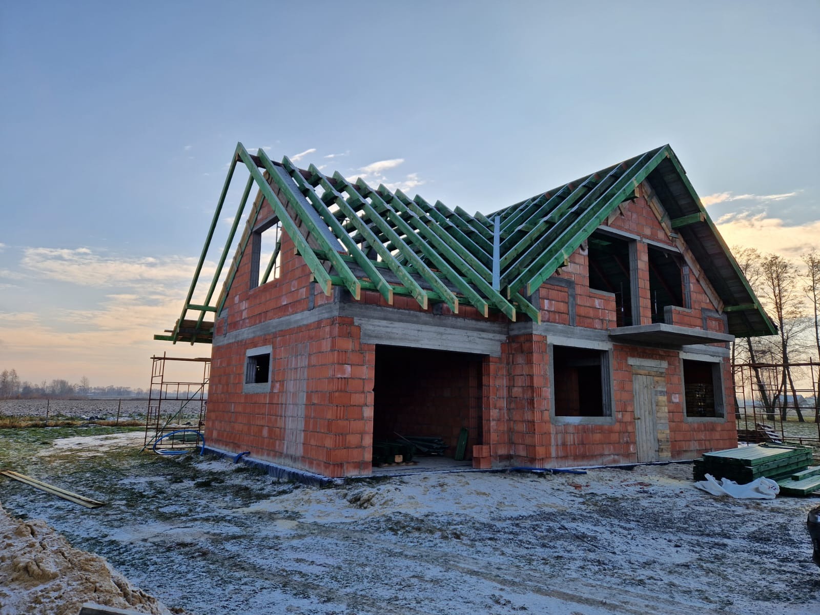 Surowy budynek jednorodzinny w stanie surowym otwartym, z czerwonej cegły, z widocznymi elementami konstrukcji dachu z impregnowanego drewna w kolorze zielonym, na częściowo ośnieżonym terenie.