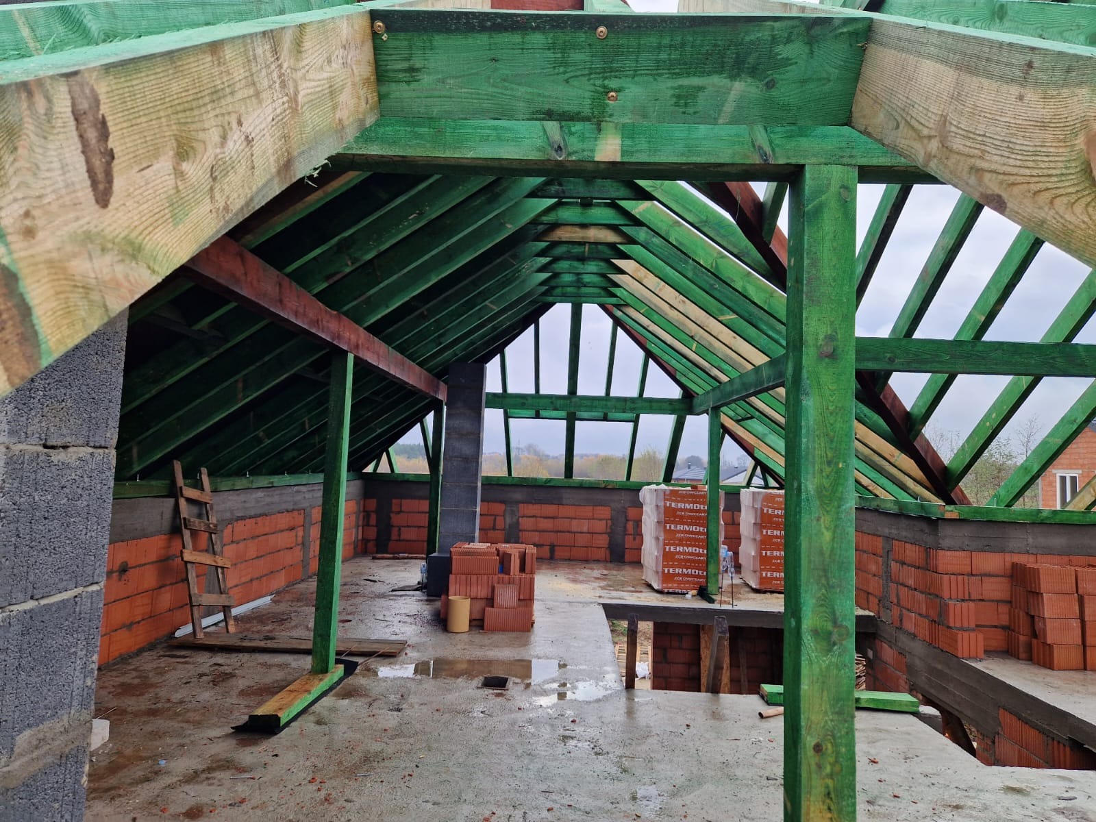 Wnętrze poddasza w trakcie budowy: widoczne ściany z czerwonej cegły, drewniana konstrukcja dachu pomalowana na zielono, palety cegieł i drabina oparta o ścianę z szarego bloczka betonowego.